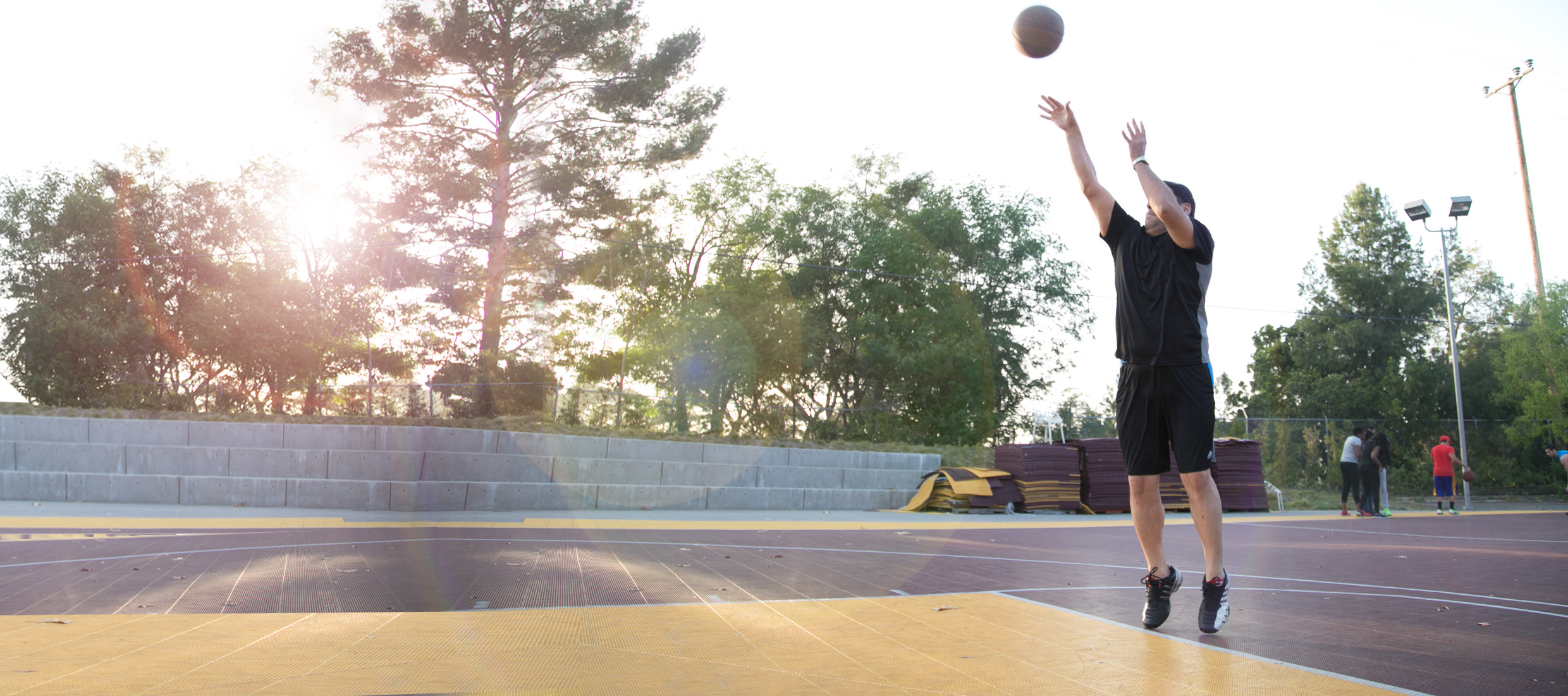 After bariatric surgery, a slim man in black throws a basketball into the air outdoors on a court in front of trees