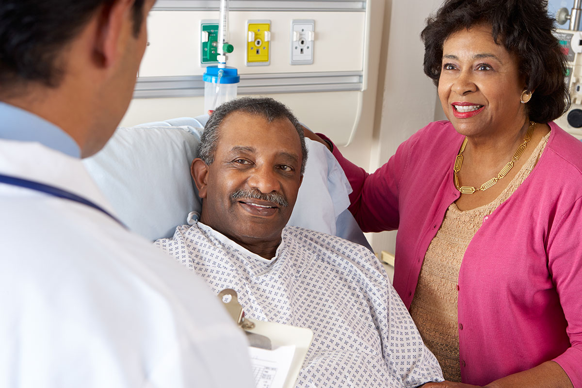 Man in a hospital bed and a woman standing bedside smile at a doctor