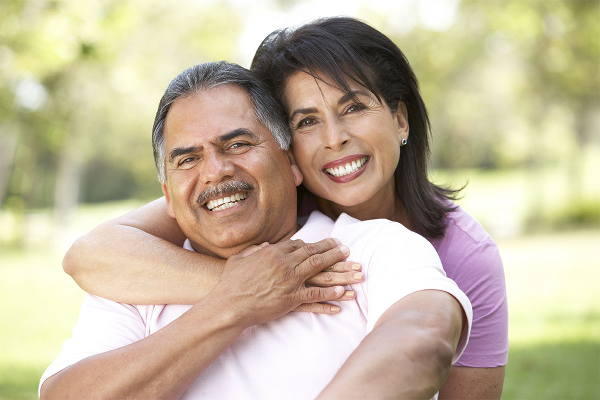 A wife and husband smile, as she hugs him around the neck from behind, and he touches her hand