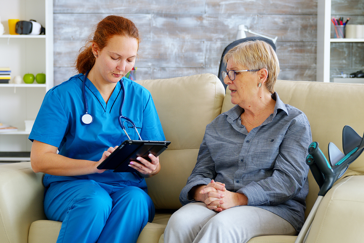 A female Keck Medicine doctor in a blue uniform, holding an iPad, interviews a female neuromuscular patient on a tan couch