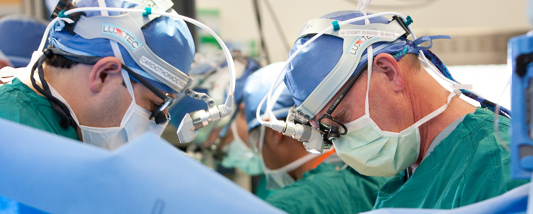 A surgeon, wearing a head lamp, glasses, mask and green scrubs, and his team perform a heart transplant in an operating room