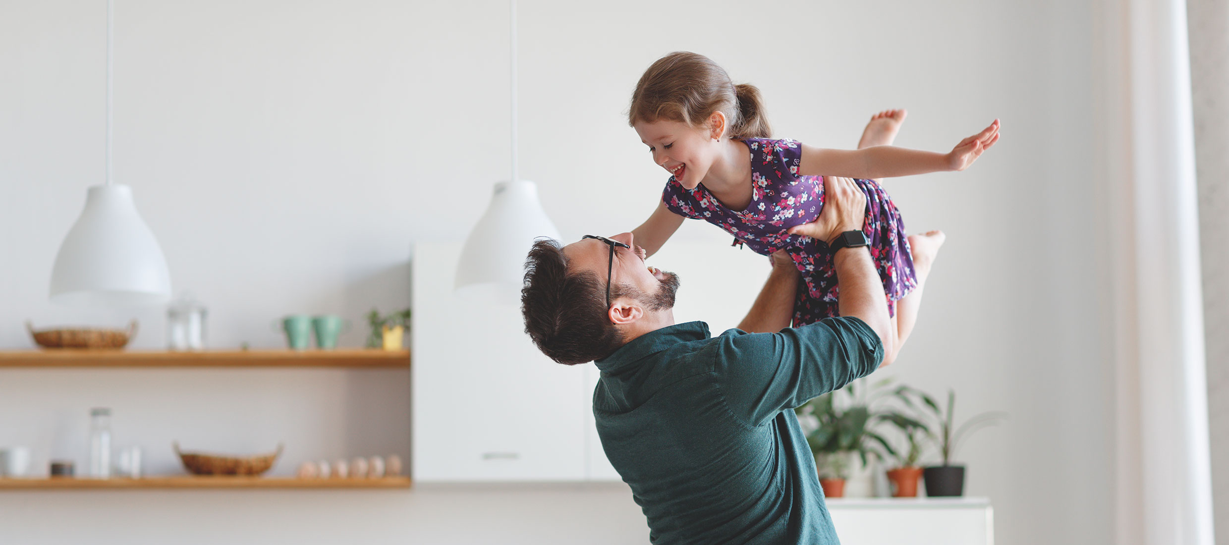 A male varicocele patient in a green shirt lifts up his smiling daughter, wearing a purple dress with her arms outstretched