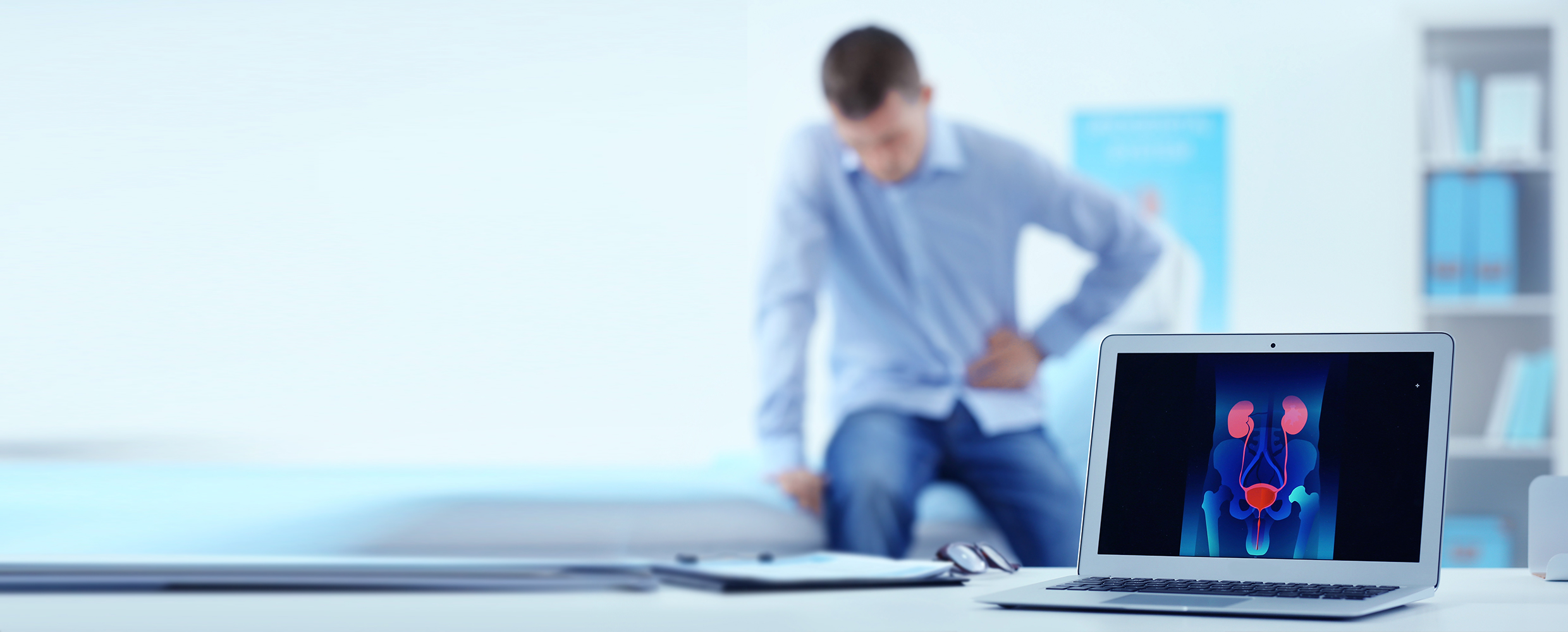 A young male urology patient in a urologist's office holds his pelvic area, and a laptop shows the male urological system