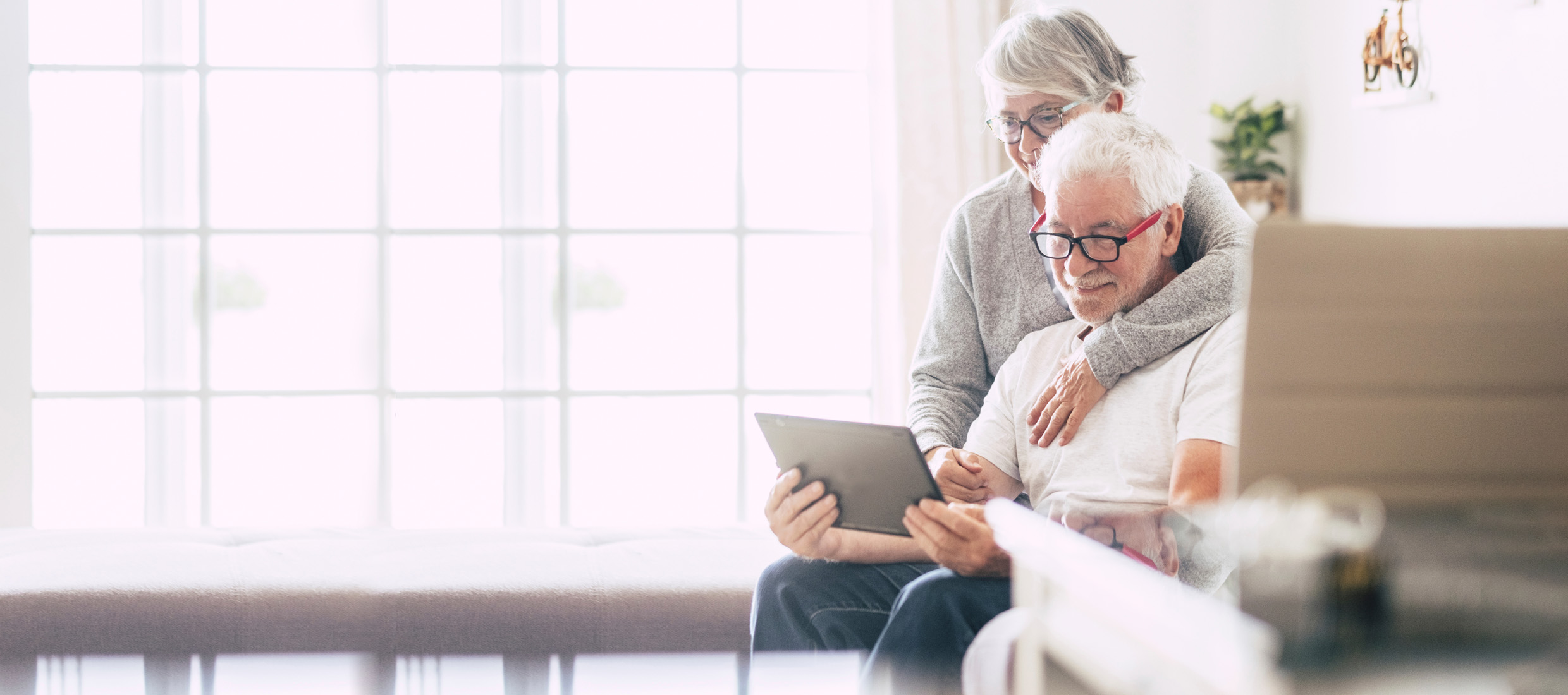 A senior male heart valve replacement patient sits on a couch with an iPad, and his wife stands behind him hugging his neck