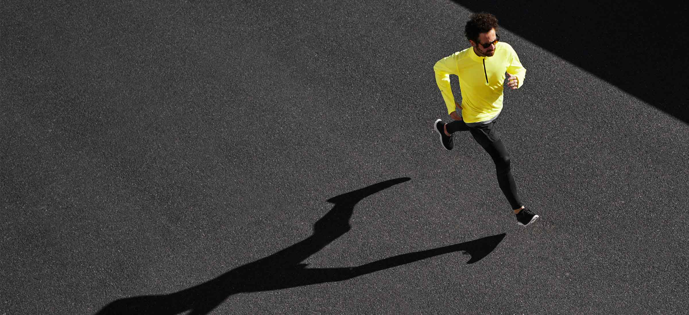 An overhead view of a man running on black pavement with his shadow.