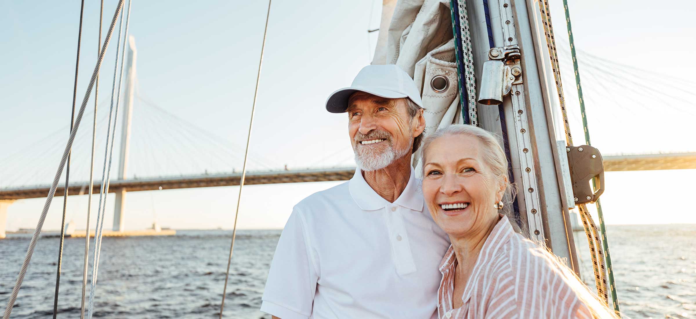 An elderly couple smiles while standing by the mast on a sailboat.
