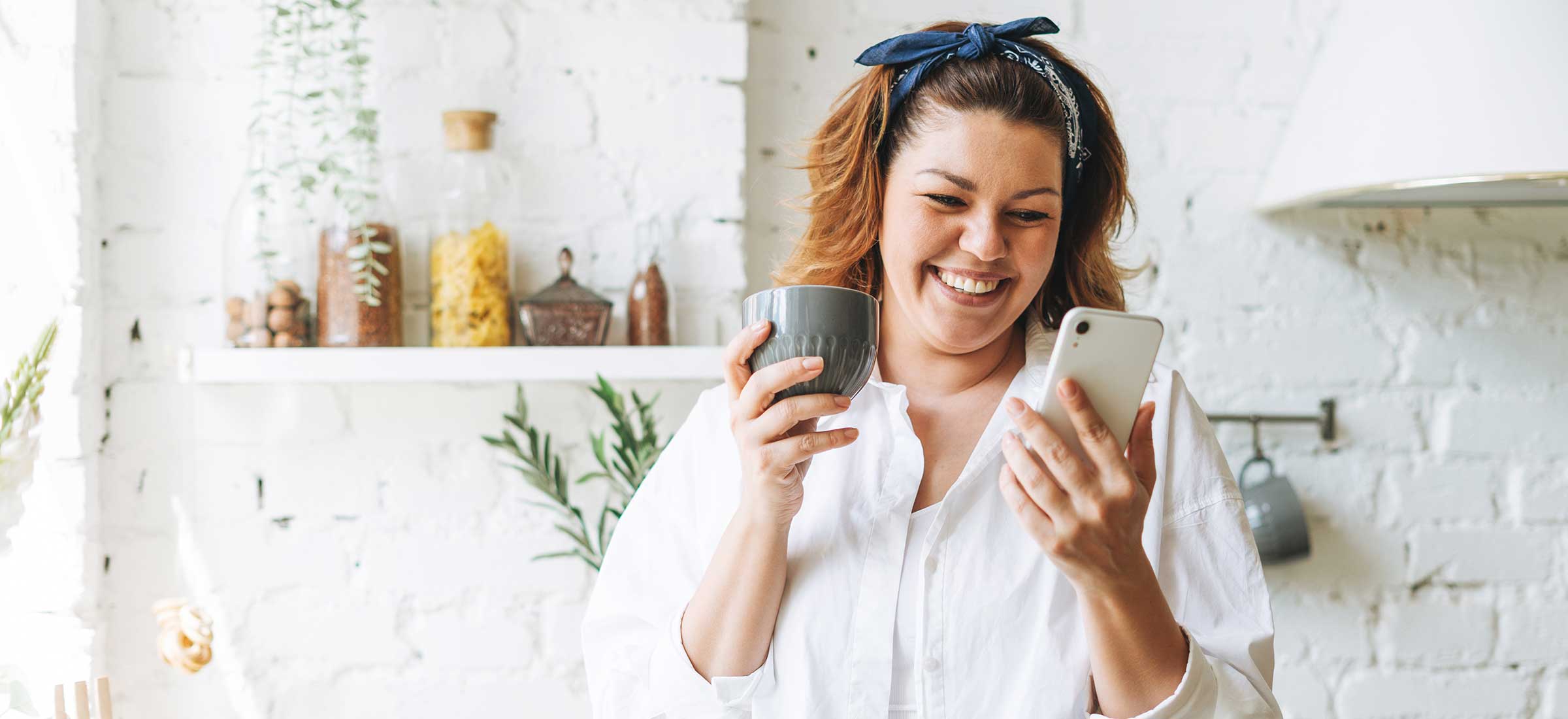 A woman smiles while looking at a cell phone and holding a coffee mug.