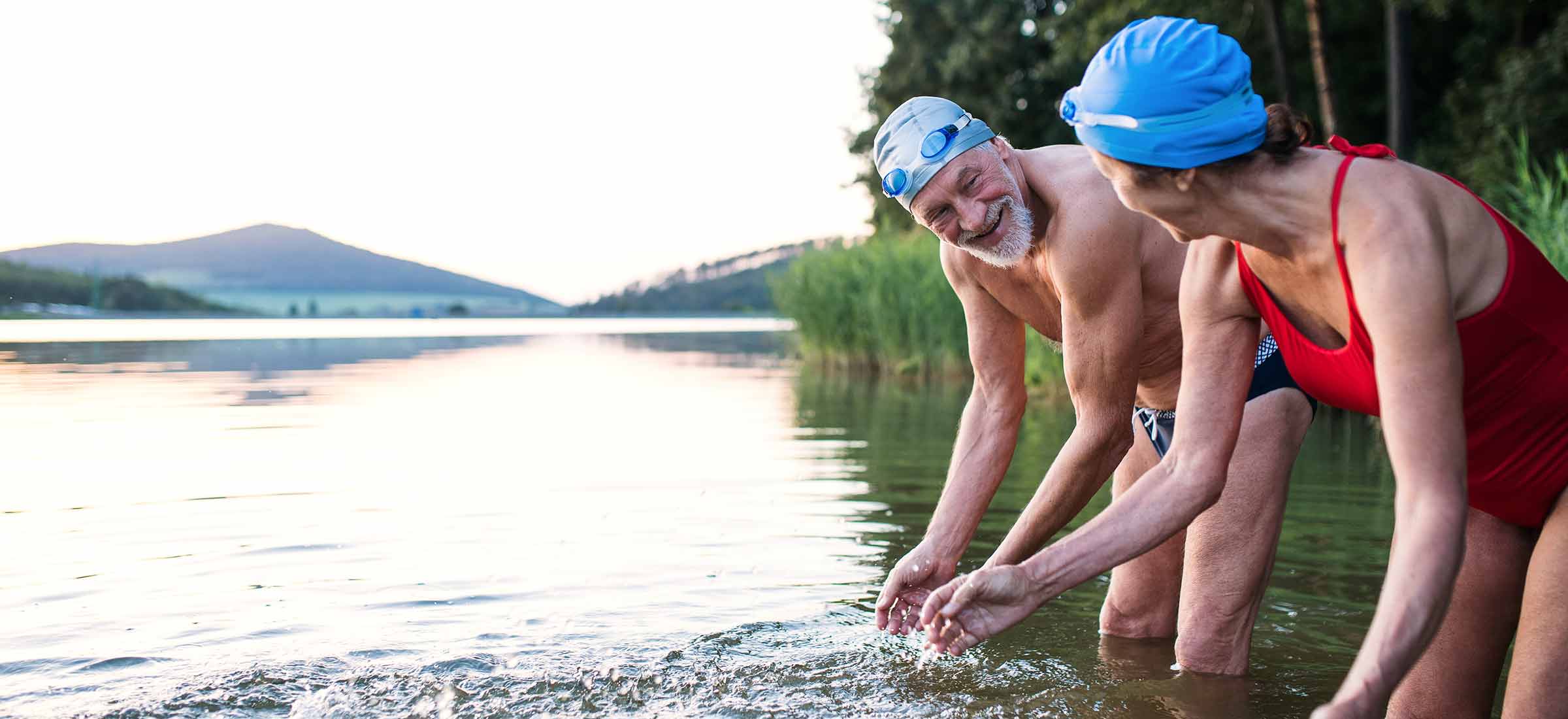 An elderly man and woman lean over in a lake with swim caps and swimsuits on.