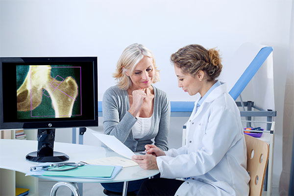A female doctor points to a paper while sitting at a table with a patient with a screen with x-ray results in the foreground.
