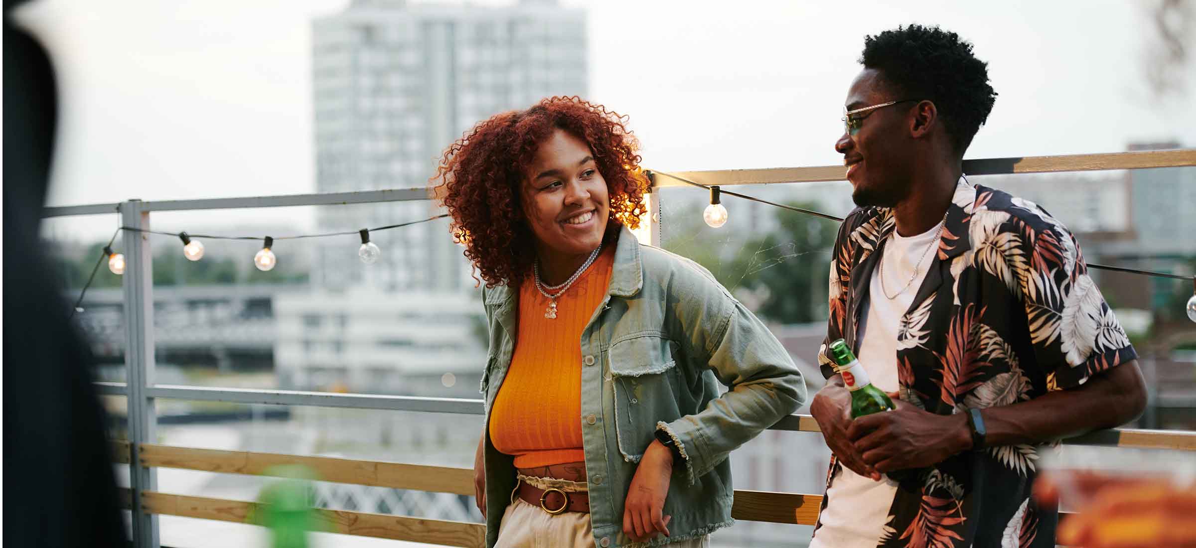 Young cheerful black woman in casualwear looking at a man during discussion on terrace of a rooftop cafe.