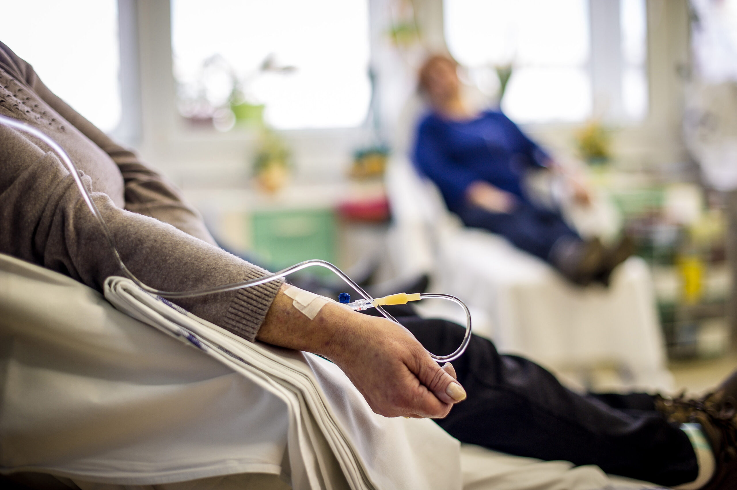 A breast cancer patient lies down in a room with another patient by windows and receives medication intravenously