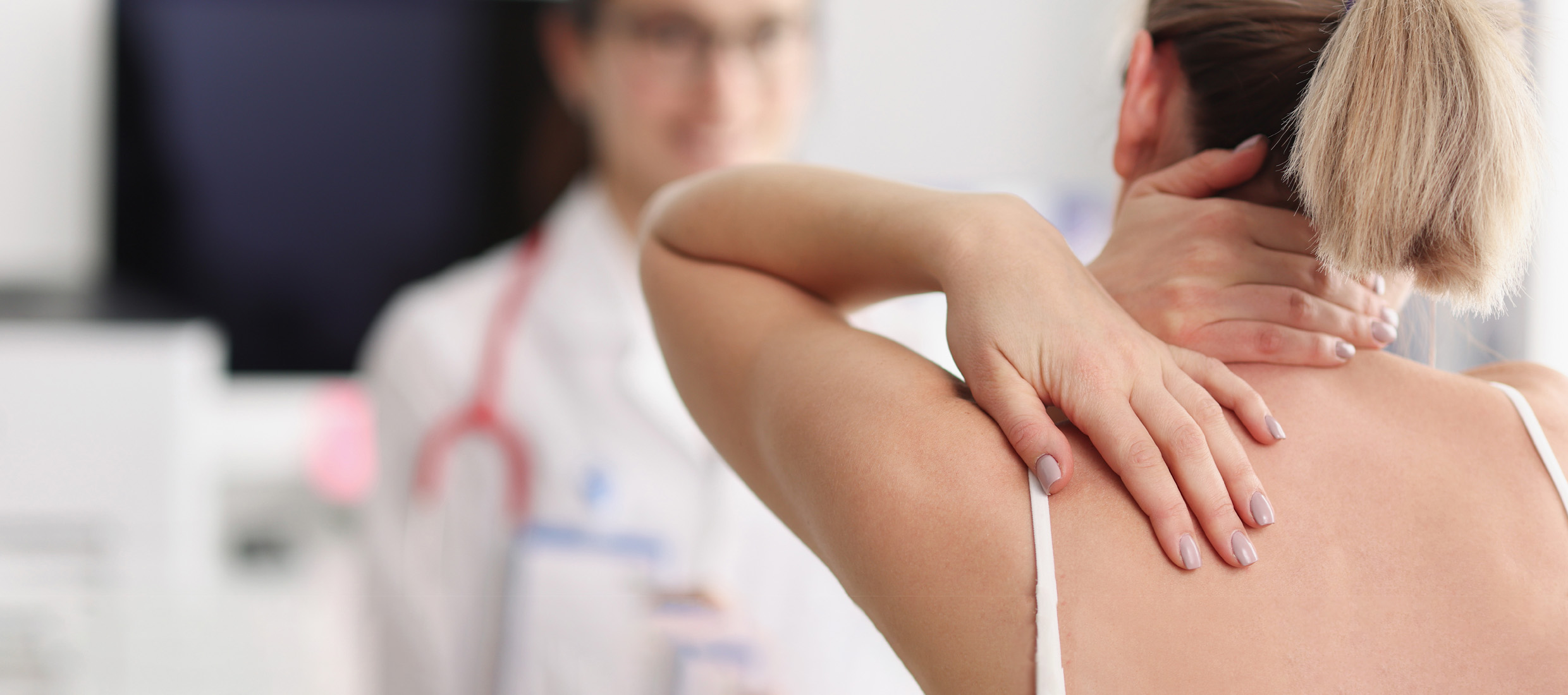 A young woman with a ponytail and neck pain holds the back of her neck and her upper back in a medical setting