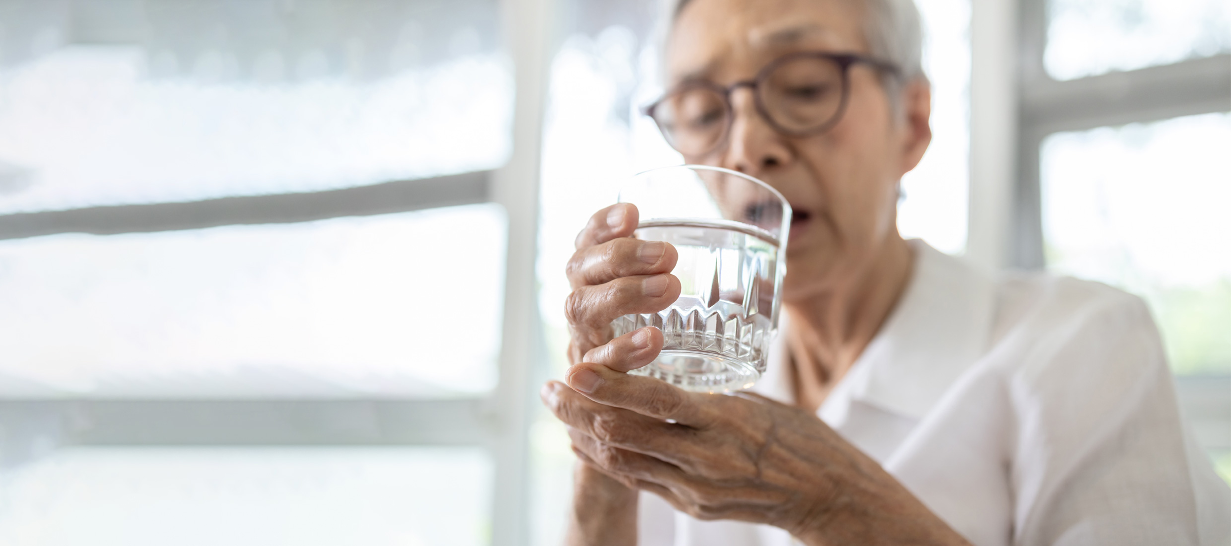 A senior Parkinson's disease patient in white shirt and glasses holds a glass of water with both hands in front of windows