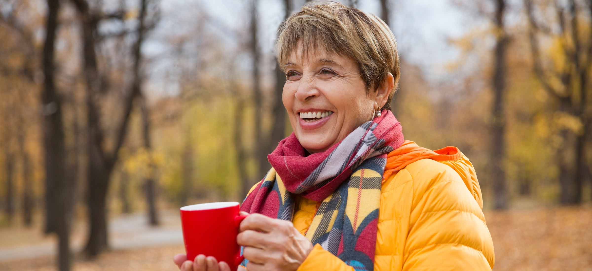 Happy senior woman in a scarf and jacket enjoying coffee at the park in autumn.