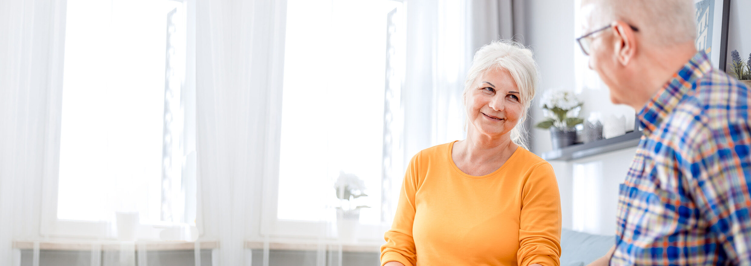 A woman and a man, both older adults, smile while sitting in a living room