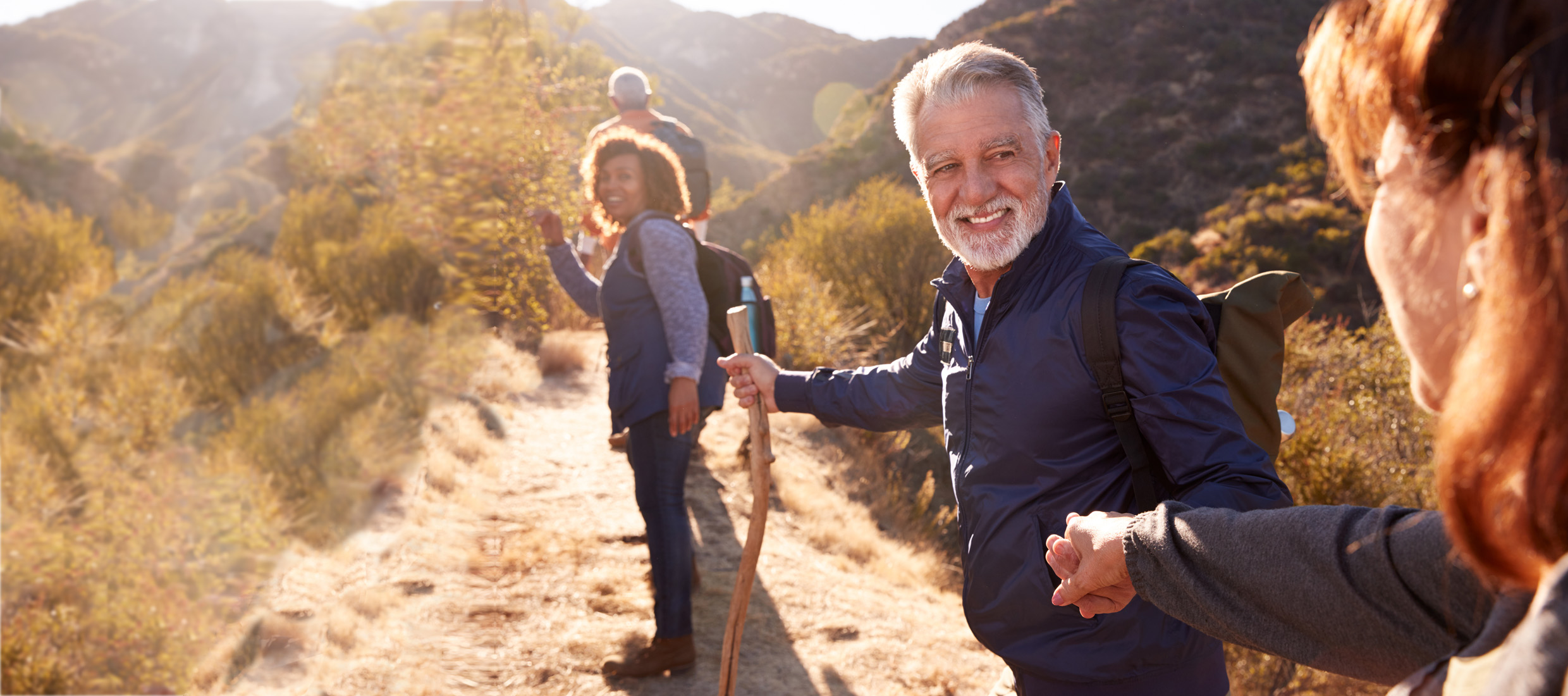 A senior man with lower back pain smiles and holds a woman's hand and uses a walking stick on a hiking trail with hikers