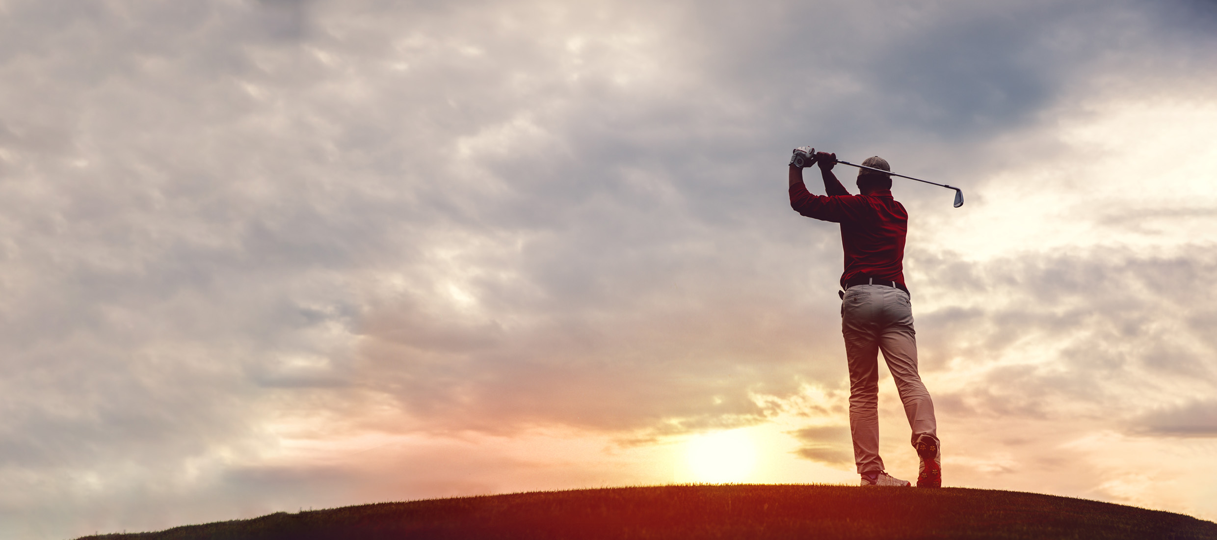 A male laminectomy patient overcomes back pain and swings a golf club on a hill in front of clouds