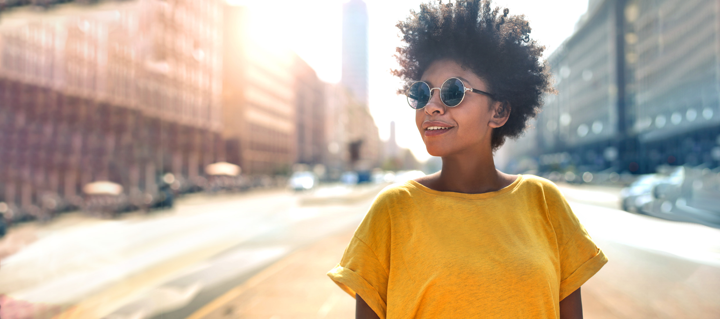 A young woman in a yellow T-shirt and round glasses walks to her dermatologist's office, smiling and looking to her right