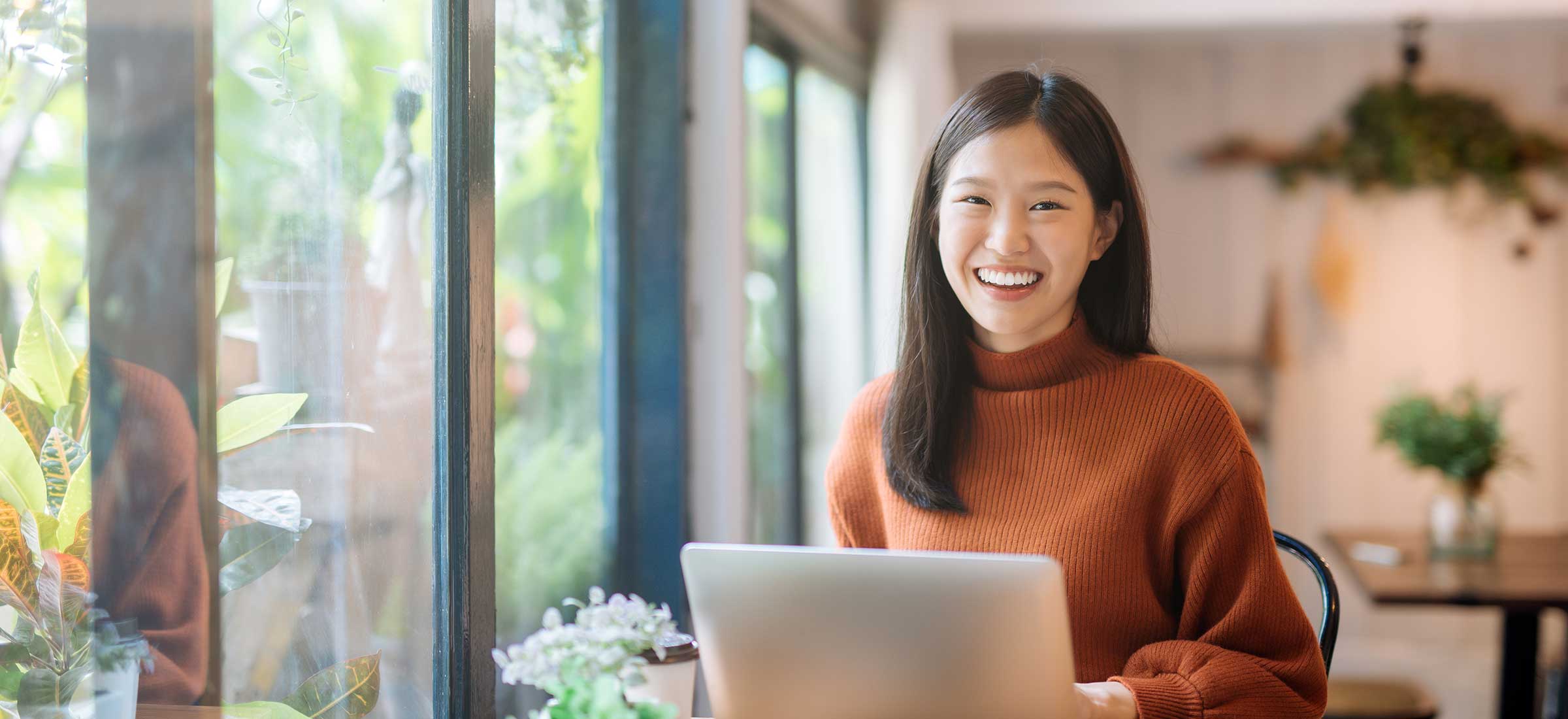 A woman sits in a room by a window on a laptop and smiles.