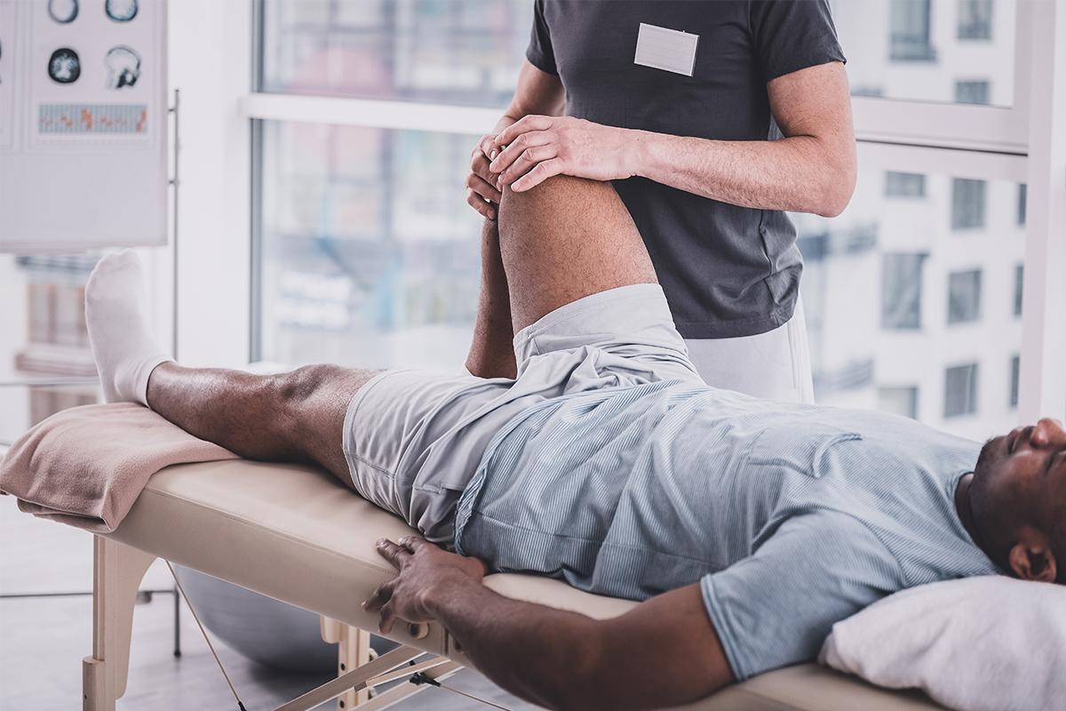 A male torn meniscus patient with knee pain lies on a table by windows and a physical therapist bends his right knee