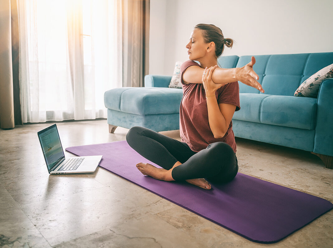 A woman seeking lower back pain relief sits cross legged on the floor, holding the elbow of her outstretched arm by a laptop