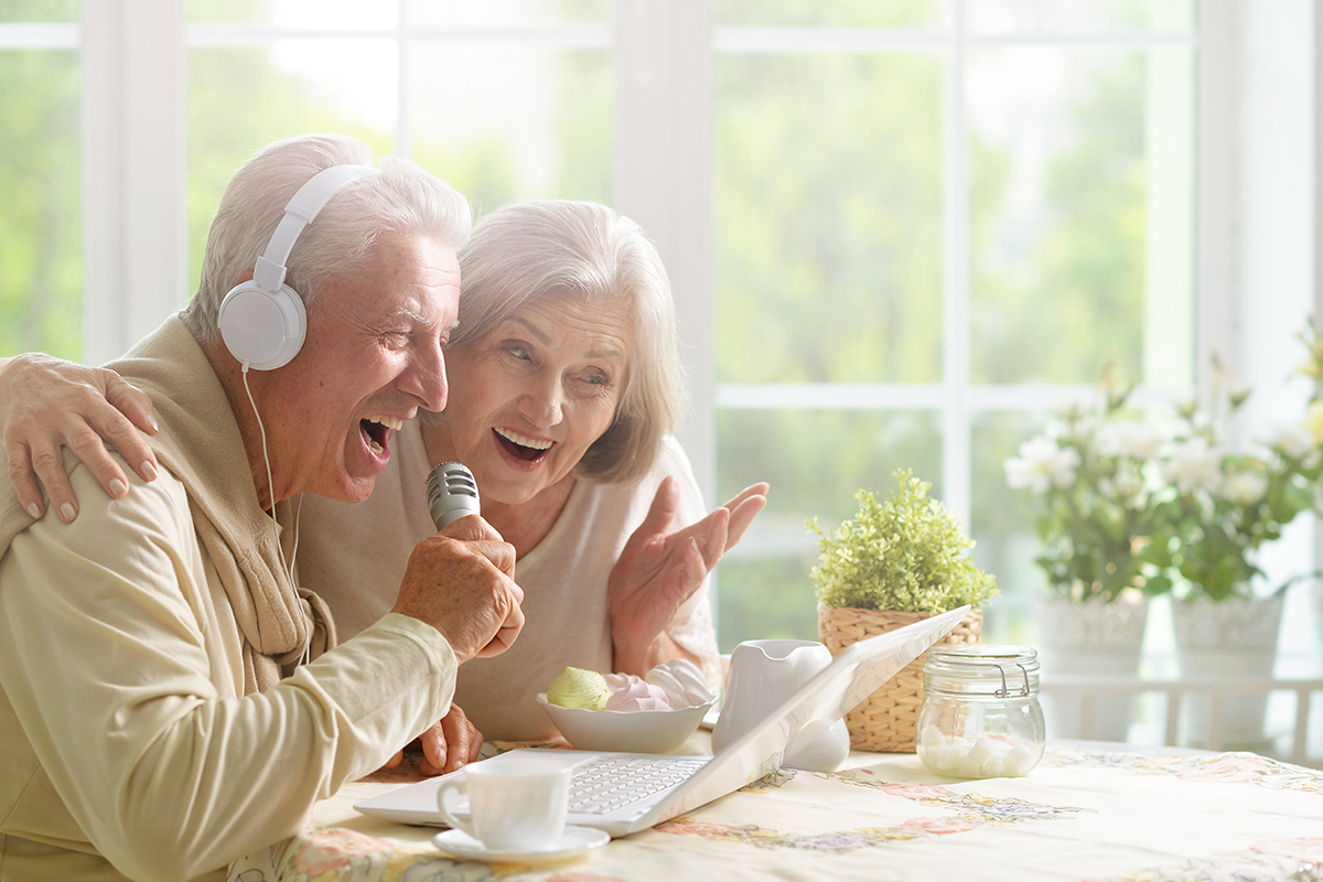 A senior sleep apnea patient wearing a headset and his wife sing into a microphone, looking at a laptop in their kitchen