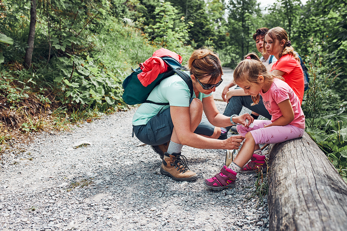 Children sit on a log on a country road, and a woman with glasses and a backpack kneels down to bandage a little girl's knee
