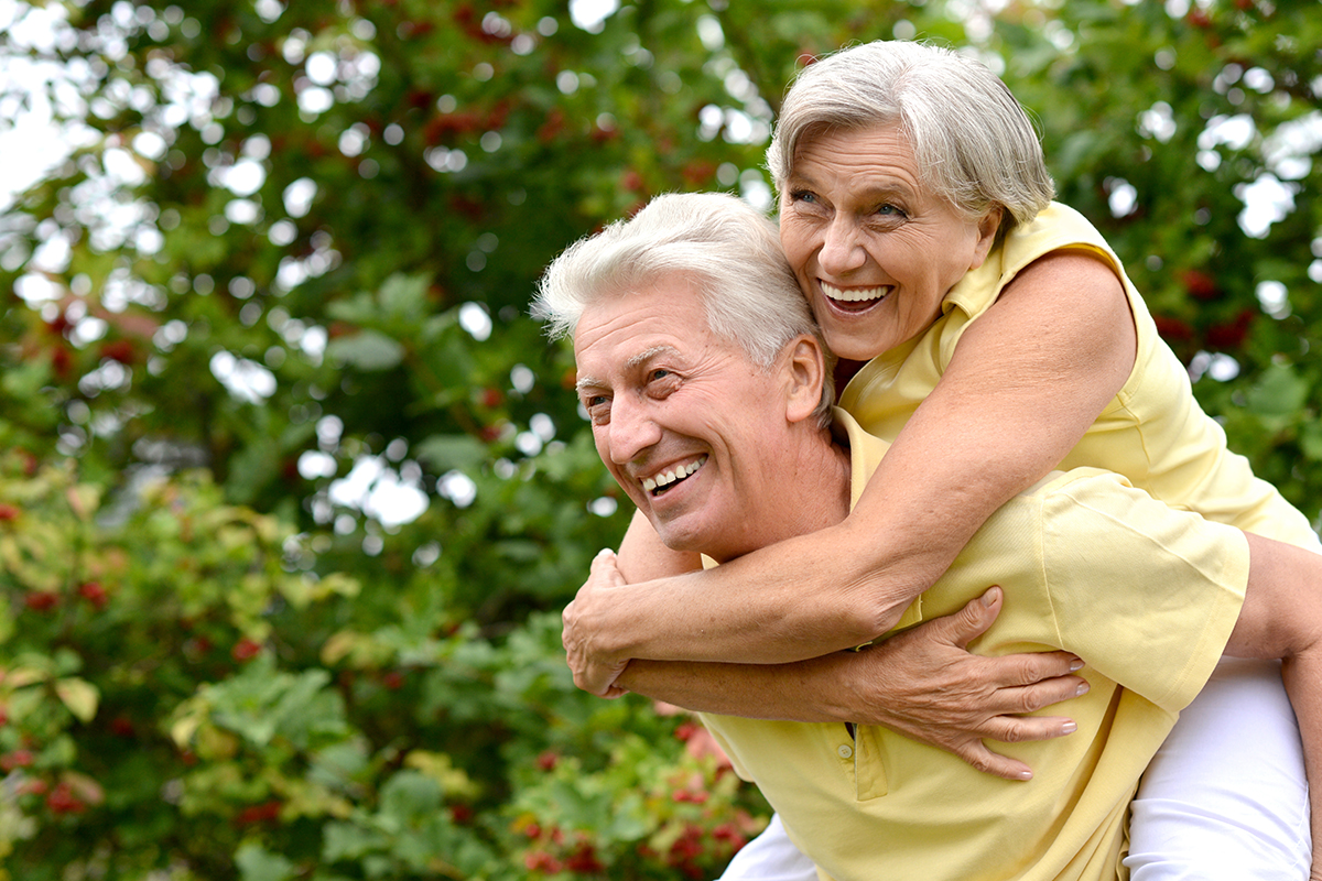 A senior male insomnia patient in a yellow shirt smiles and gives a senior woman a piggy back ride in front of trees