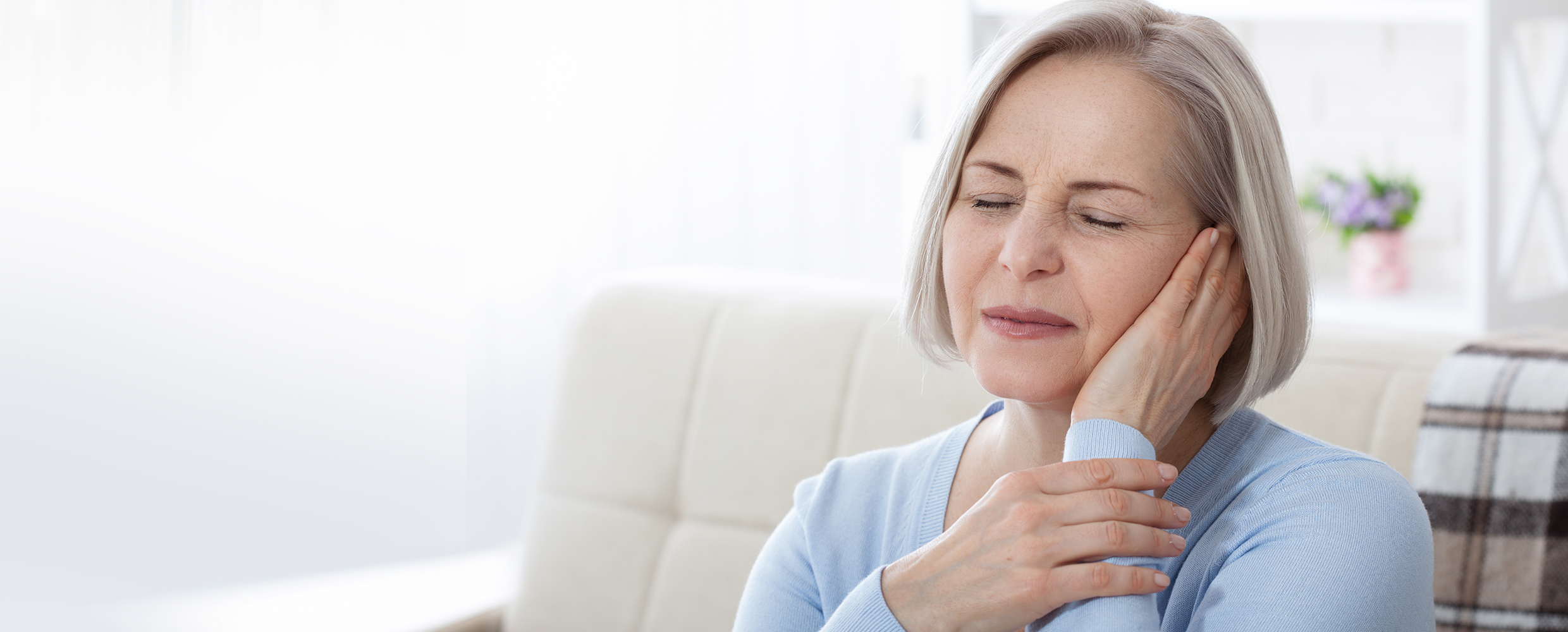 A senior trigeminal neuralgia patient, wearing a blue sweater, grimaces with pain and holds her left hand to her left ear