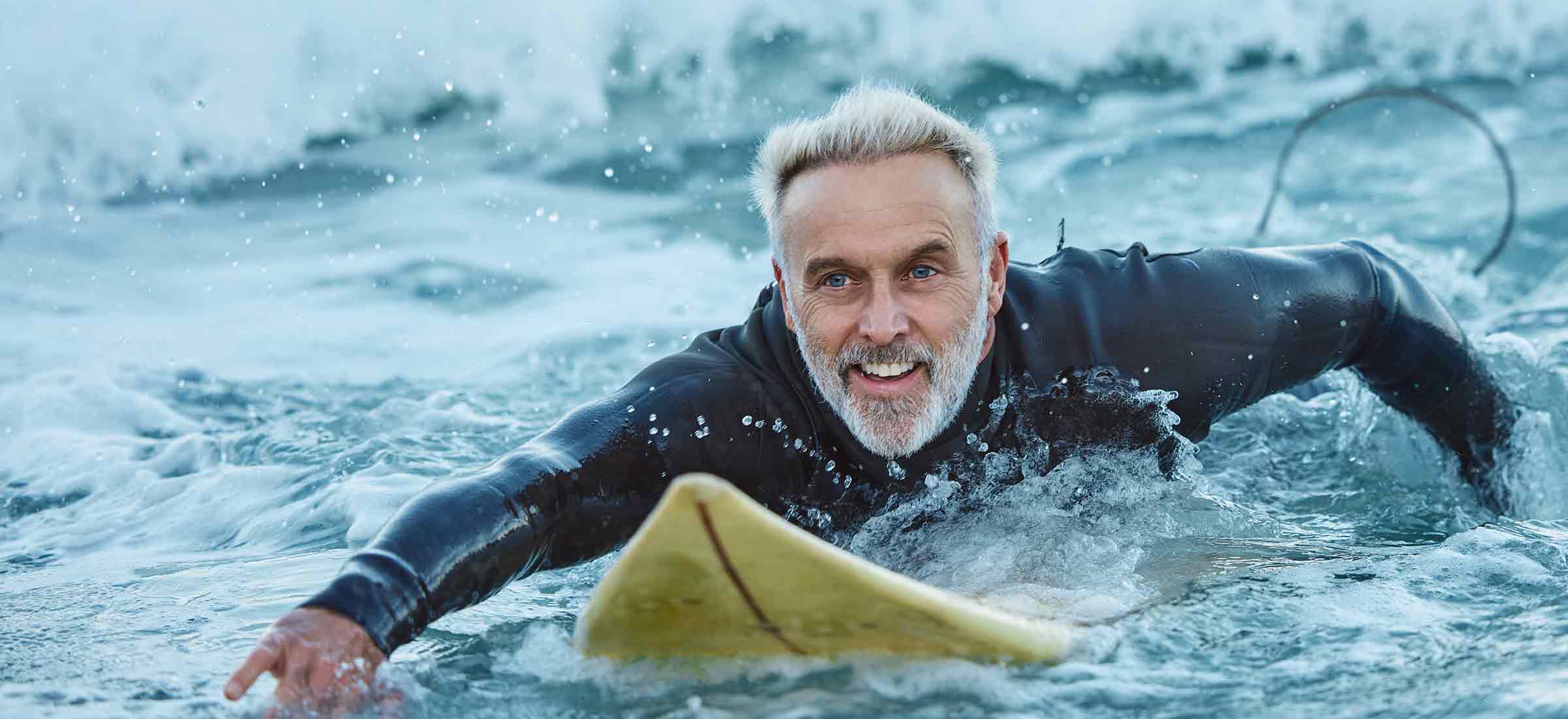A senior man paddles through waves on a surfboard in the ocean.