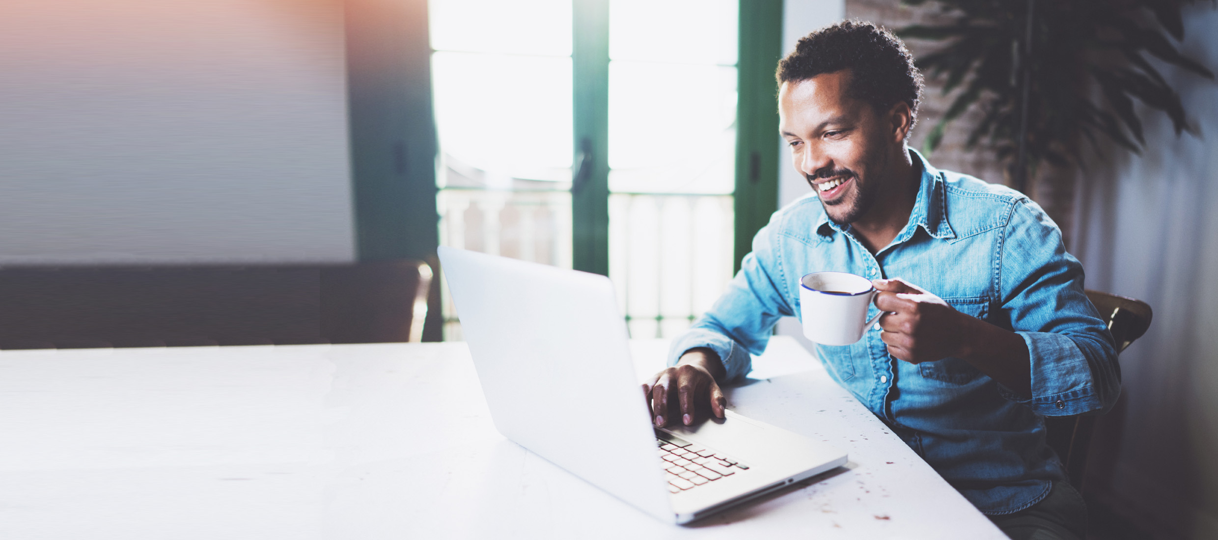 A male LASIK eye surgery patient in a denim shirt holds a coffee cup and uses a laptop in front of windows