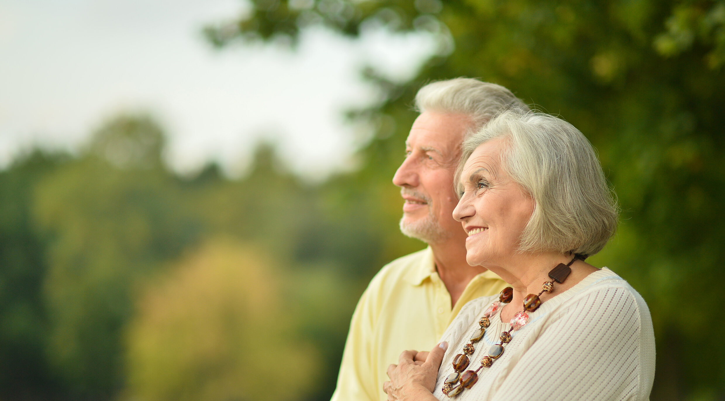 A senior female lung cancer patient, wearing a necklace, and her husband smile and look up at the sky in the park