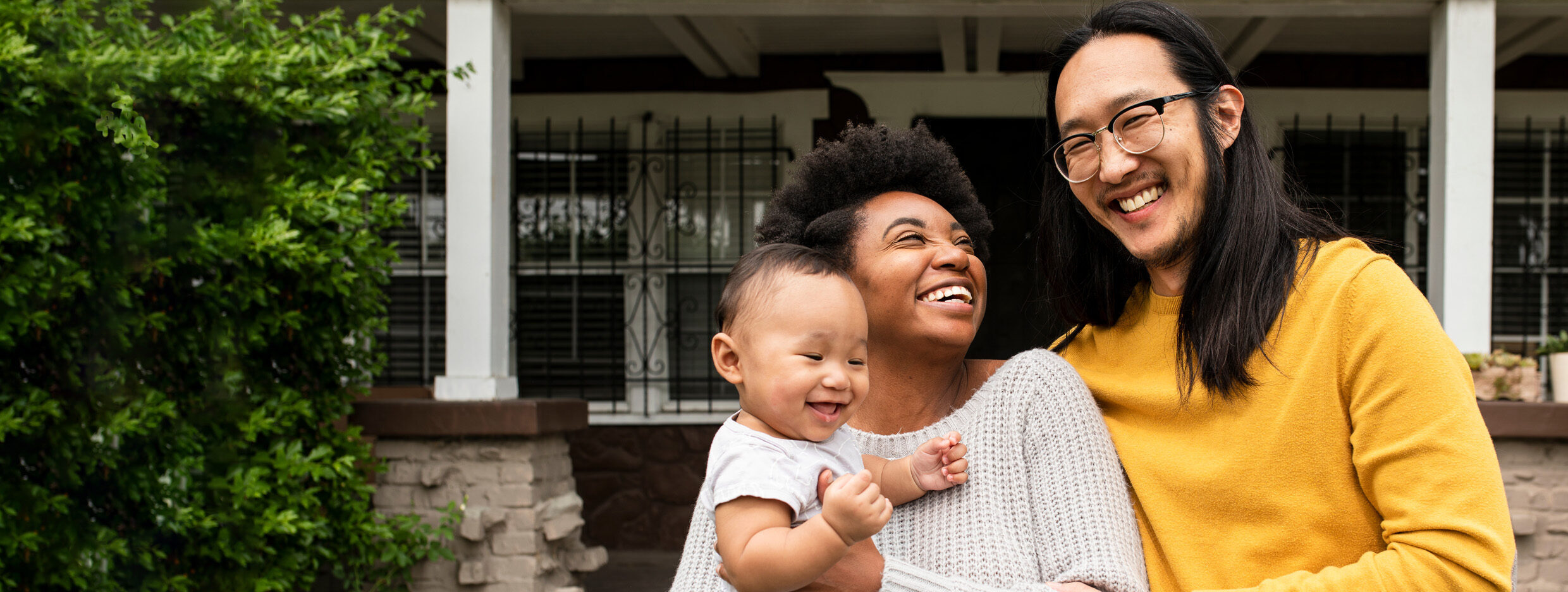 A man and a woman holding an infant smile while standing in front of a house