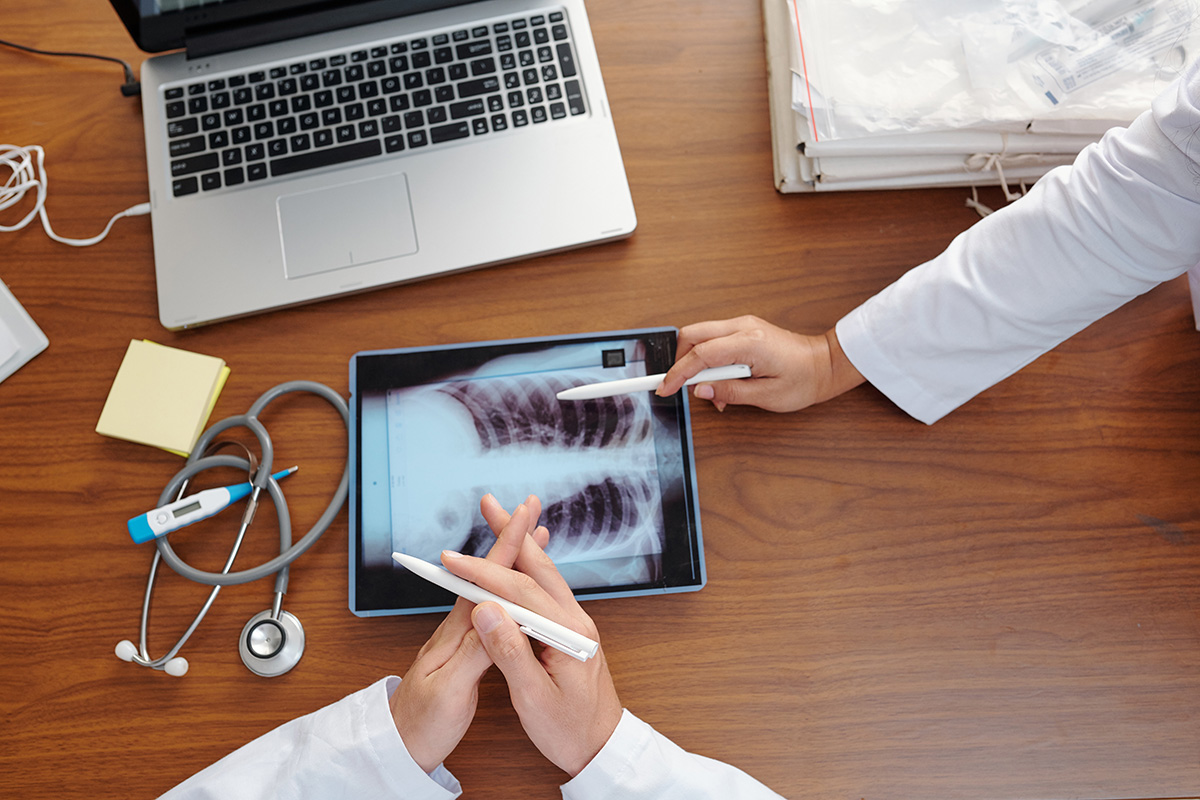 Two Keck Medicine pulmonologists point to chest X-rays on an iPad on a wood table with a laptop, stethoscope and thermometer