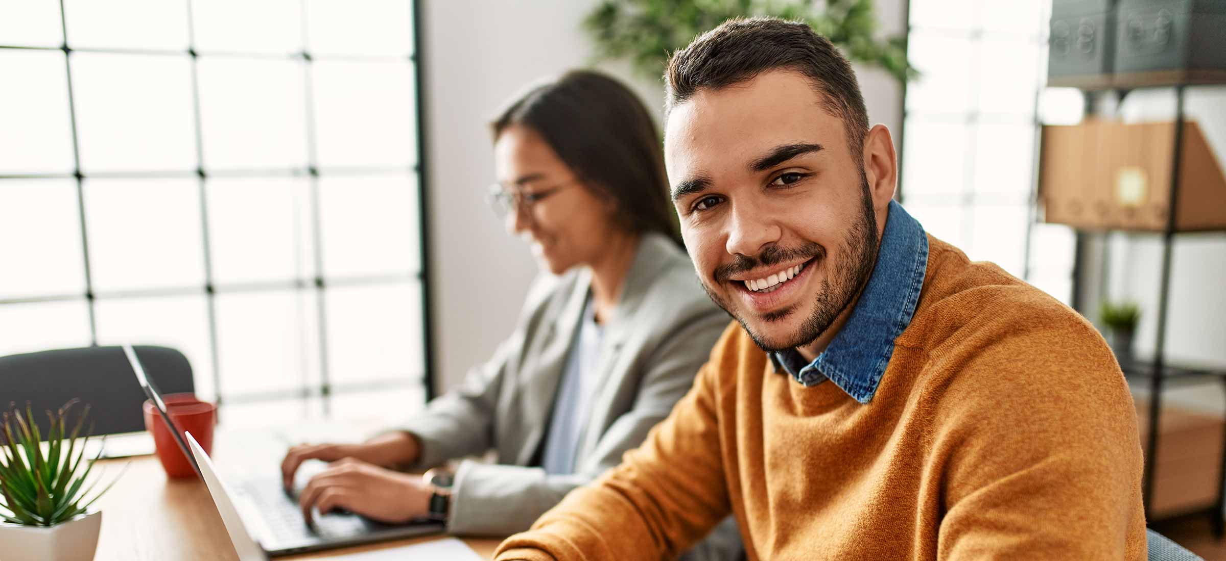 Two coworkers smiling happily while sitting at their desks in the office.