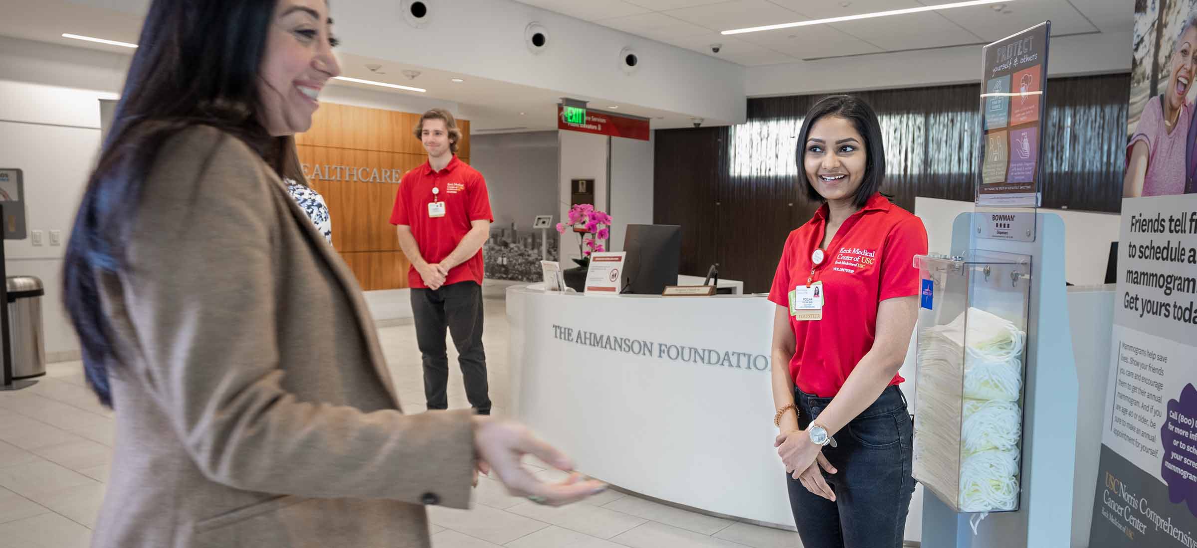 two hospital volunteers stand at the entrance to Keck Medicine of USC and greet a patient