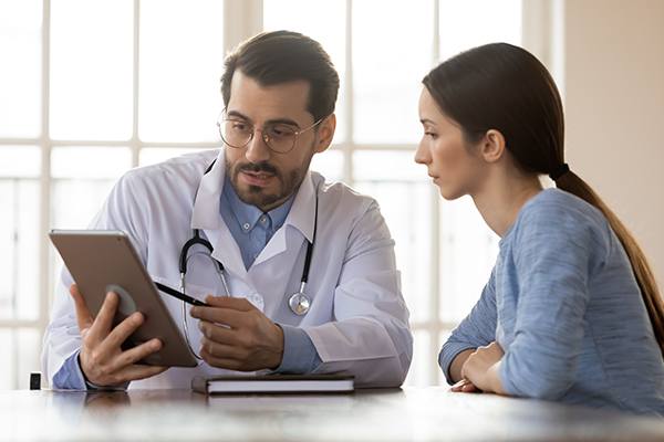 Focused young male doctor pointing at digital computer tablet screen, sharing health tests laboratory results to serious female patient.