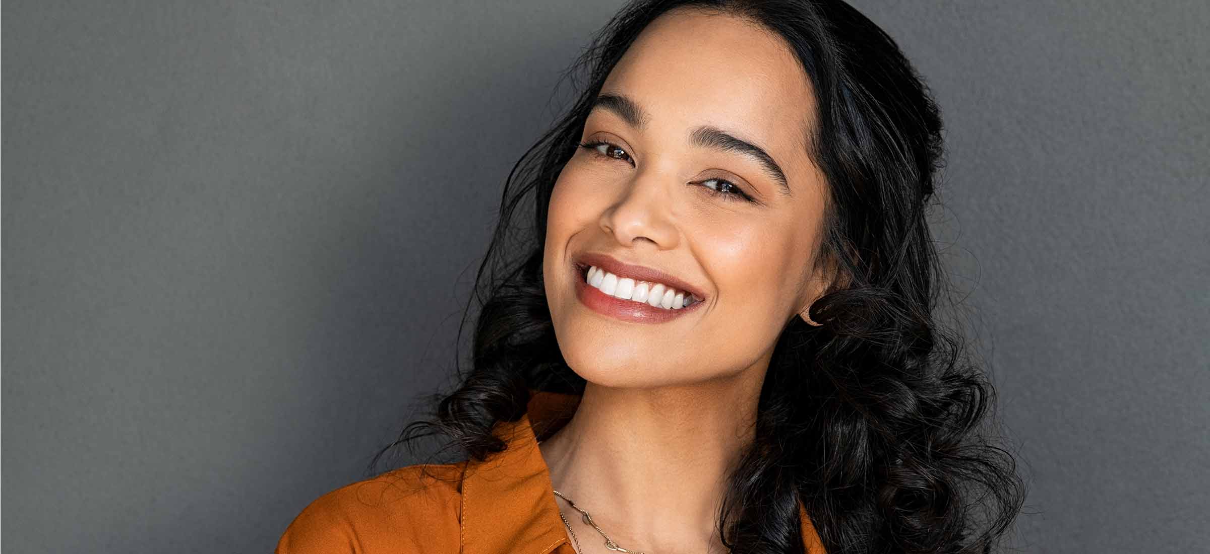 Latin woman looking at camera against a gray wall with a big smile.
