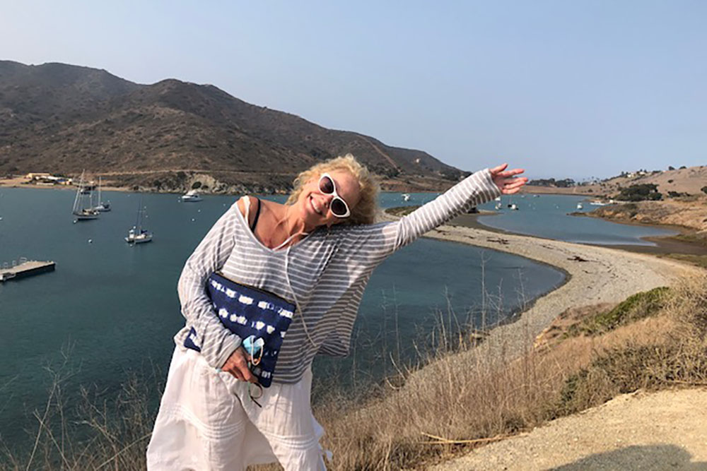 Actress DeeDee Rescher smiles and waves her arm on a hillside in front of a pristine harbor