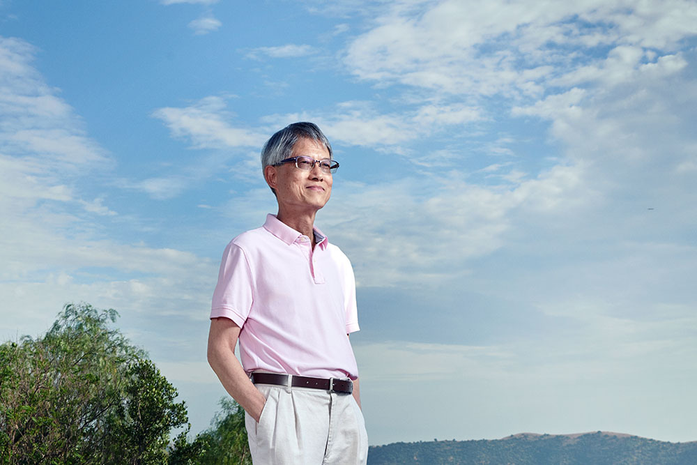 Danny Kwok, an older Asian-American man, stands with his hands in his pockets, wearing a pink shirt tucked into tan slacks