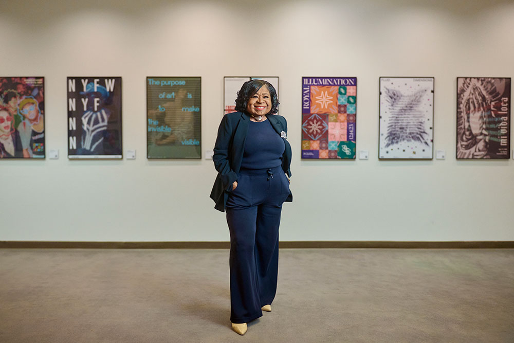 Cora Jackson-Fossett, a Black woman in a blue suit, smiles in front of an exhibit of paintings