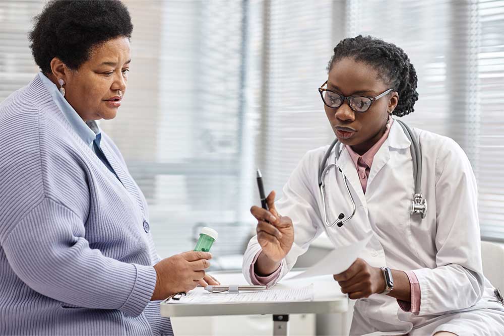 A doctor looks at a paper on a table in front of a patient holding a bottle of pills.