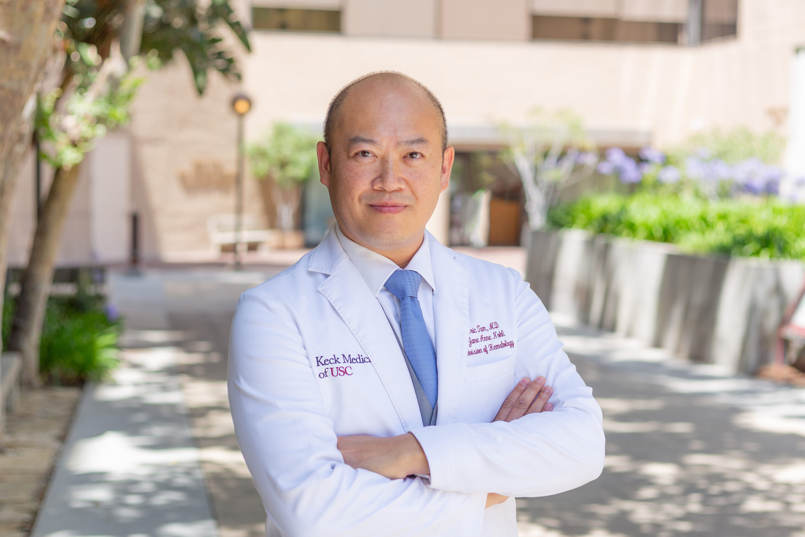 A portrait of Dr. Eric Leon Tam standing outside in a white medical coat crossing his arms.