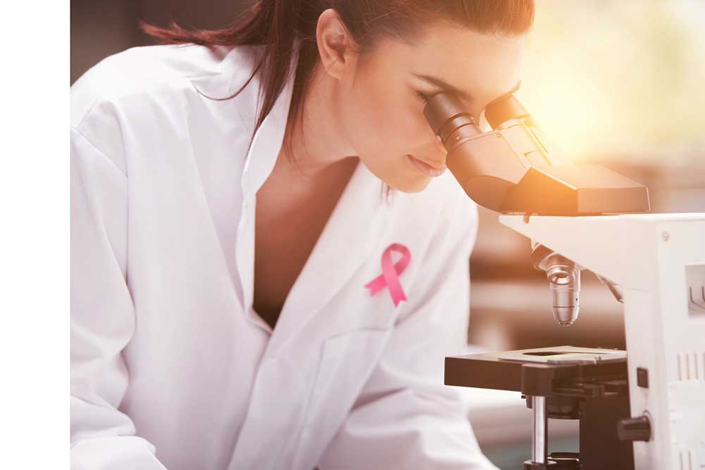 Researcher with breast cancer awareness emblem on white coat looks through microscope.