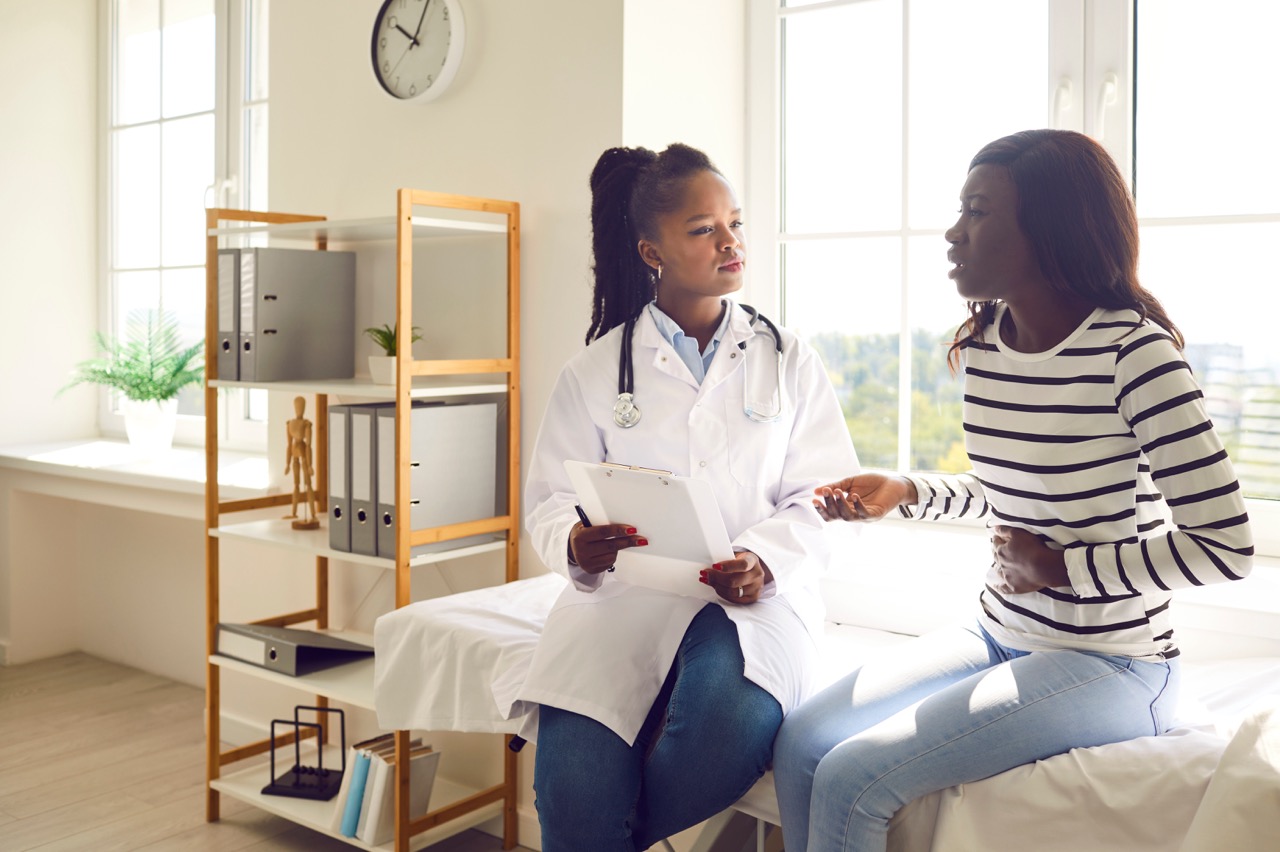 Young sick woman with abdomen pain visiting her female therapist doctor in medical clinic