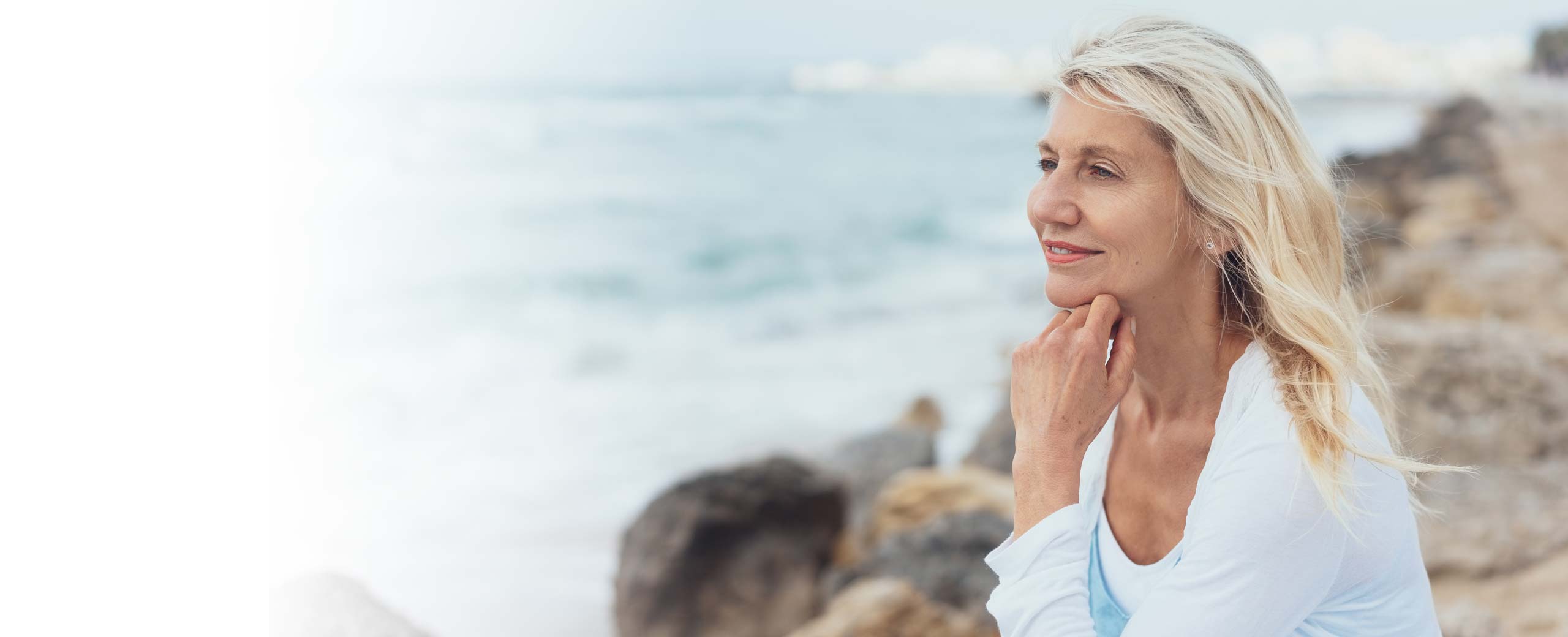 Senior woman relaxes while sitting on the rocks next to ocean.