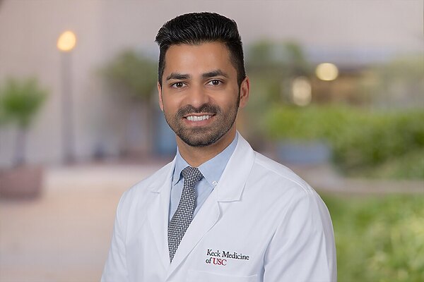 Dr. Sij Hemal stands in front of a blurred background in a white medical coat, smiling.
